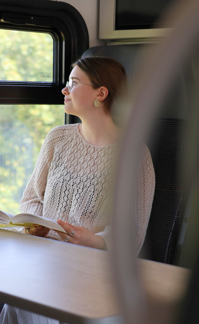 Junge Frau sitzt im Zug, schaut aus dem Fenster und hat ein geöffnetes Buch in den Händen